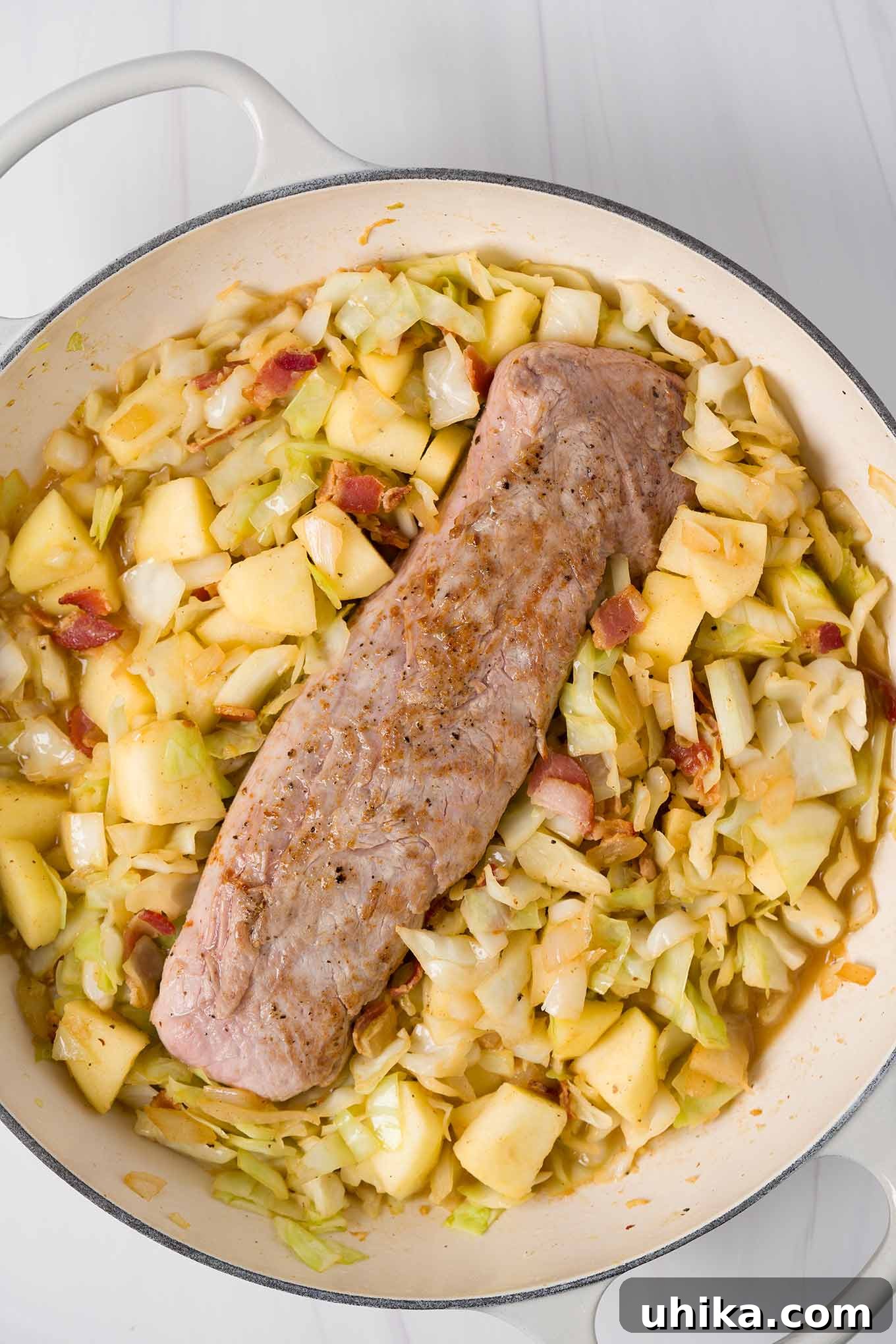 A preparation photo for braised pork tenderloin showing the pan-seared pork ready for braising with the apples, cabbage, bacon, and apple cider.