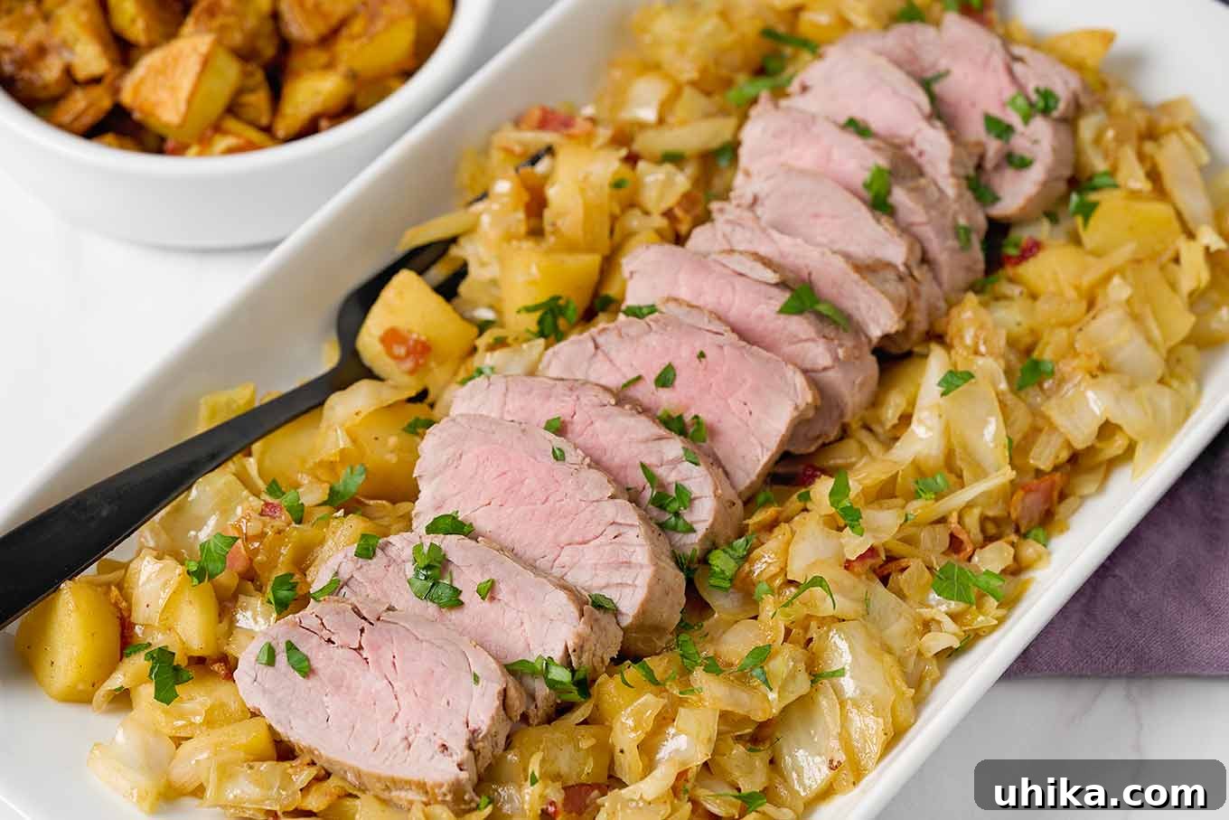 A white serving platter of cider-braised pork tenderloin with cabbage and apples, with a plum-colored napkin and dish of roasted potatoes to the side.