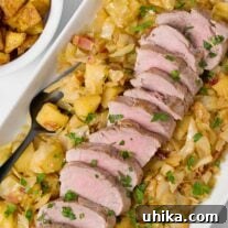 A platter of braised pork tenderloin, sliced and served on a bed of cabbage, apples, and bacon, a bowl of roasted potatoes on the side.