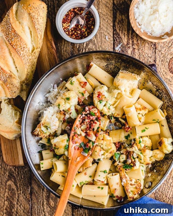 Rigatoni with Cauliflower and Bacon - Hearty Italian Dinner A large pan filled with Rigatoni with cauliflower and bacon, surrounded by grated cheese, crusty bread, and chili flakes on a wooden table.