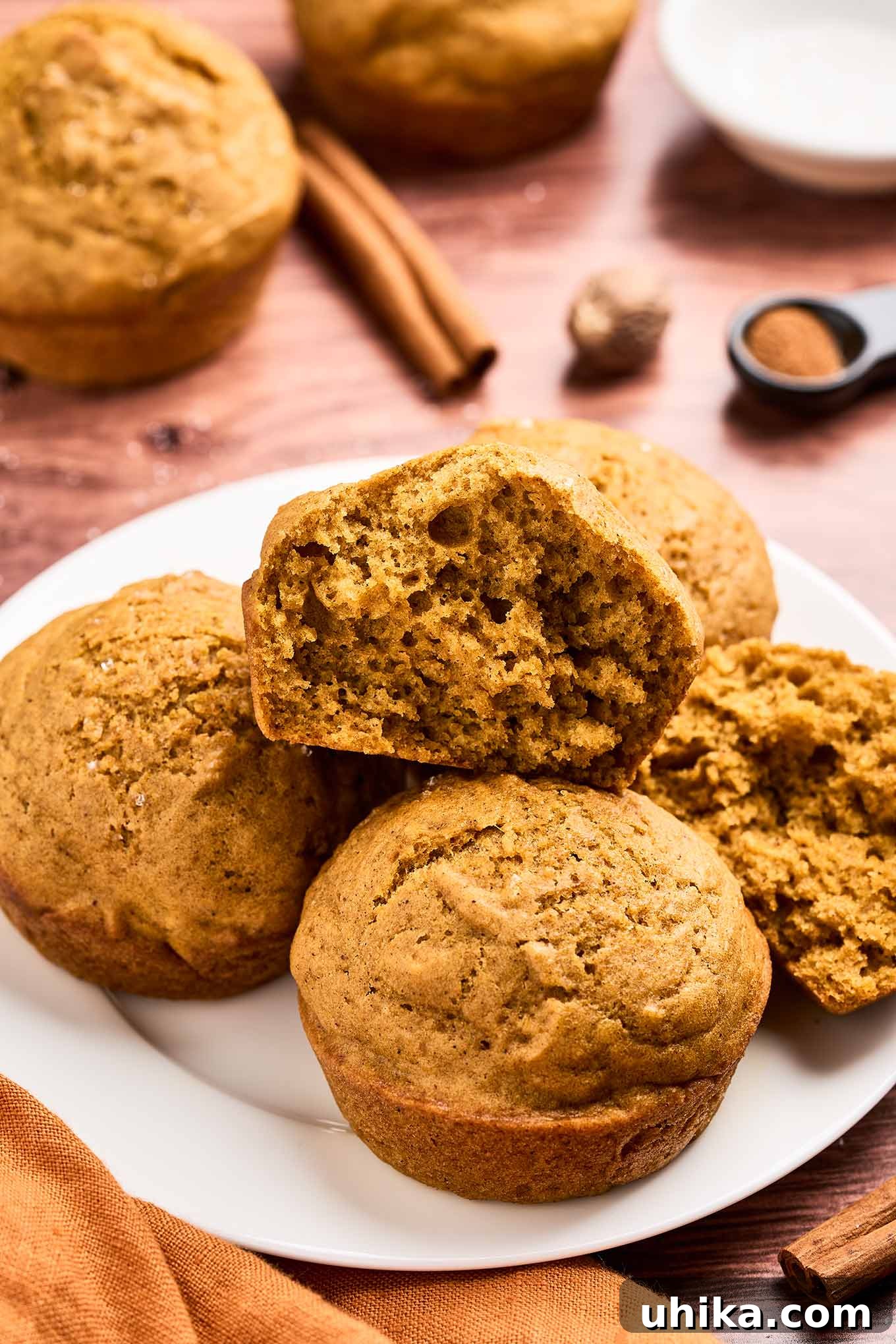 Cozy Pumpkin Spice Bites 2 Close-up of moist and fluffy pumpkin spice muffins on a plate, one cut open to show soft texture, cinnamon sticks in the background.