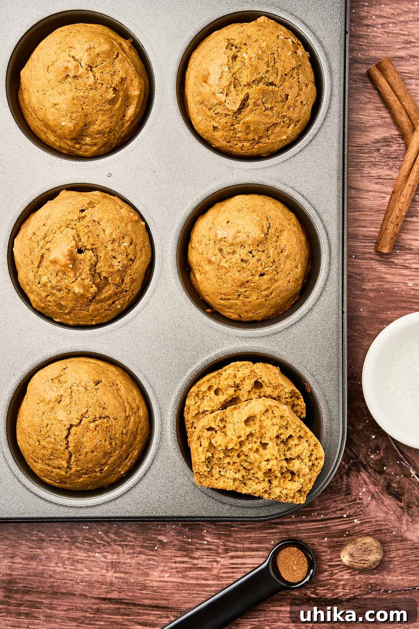 Cozy Pumpkin Spice Bites 6 Pumpkin spice muffins with golden brown tops, cooling in a muffin tin, surrounded by cinnamon sticks, whole nutmeg, and spoon of ground cloves.