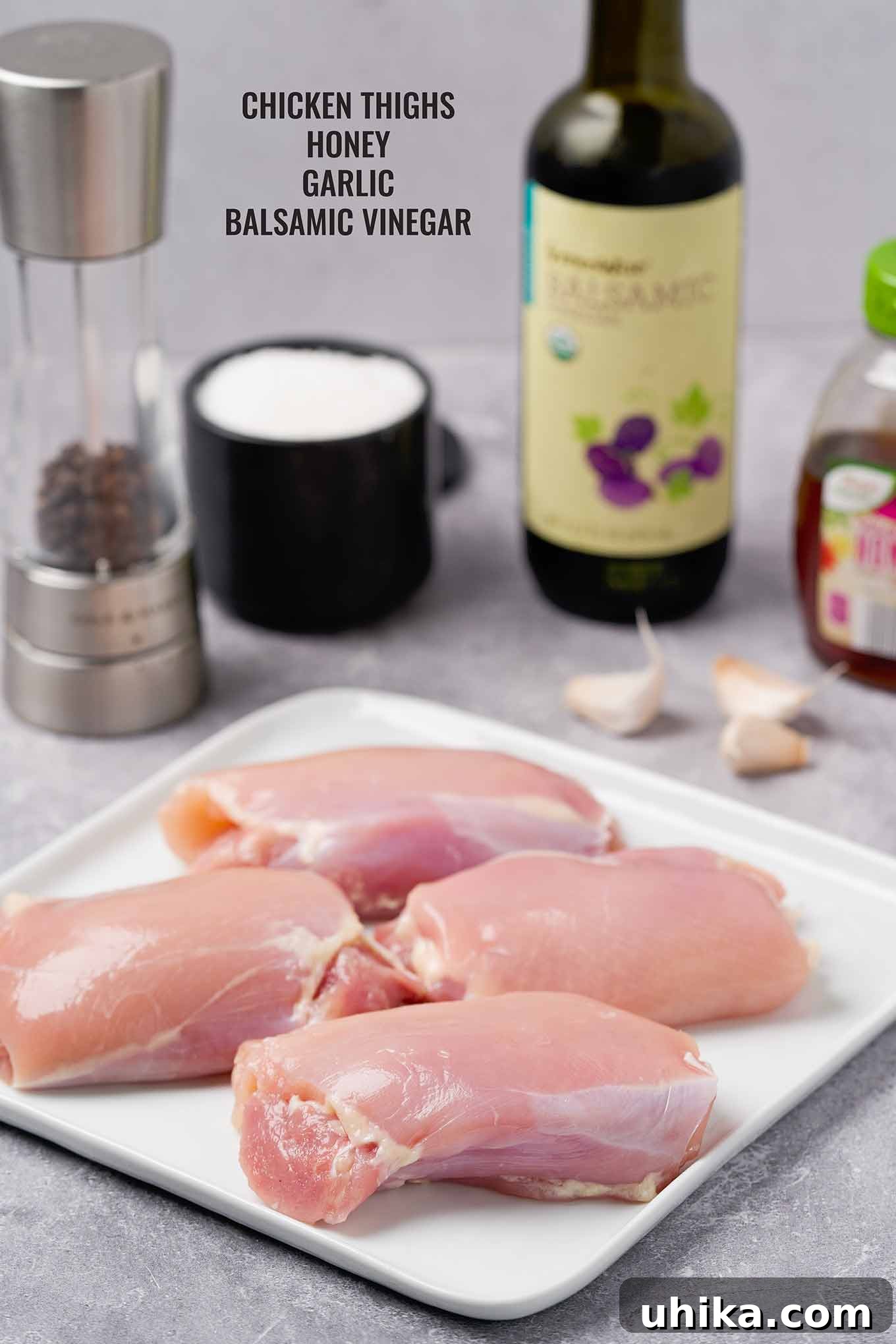 Succulent Oven-Roasted Boneless Chicken Thighs 3 Labeled ingredient photo focused on boneless, skinless, chicken thighs with balsamic vinegar, honey, garlic cloves, and salt and pepper in background.