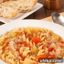 A bowl of chicken soup with curly noodles, chicken, carrots, celery, tomatoes, and cabbage, surrounded by a napkin, spoon, peppermill, and plate with crusty bread.