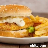 An oven-fried fish fillet on a seeded soft roll with melted cheddar cheese, tartar sauce, and lettuce, and fries on the plate to the right.