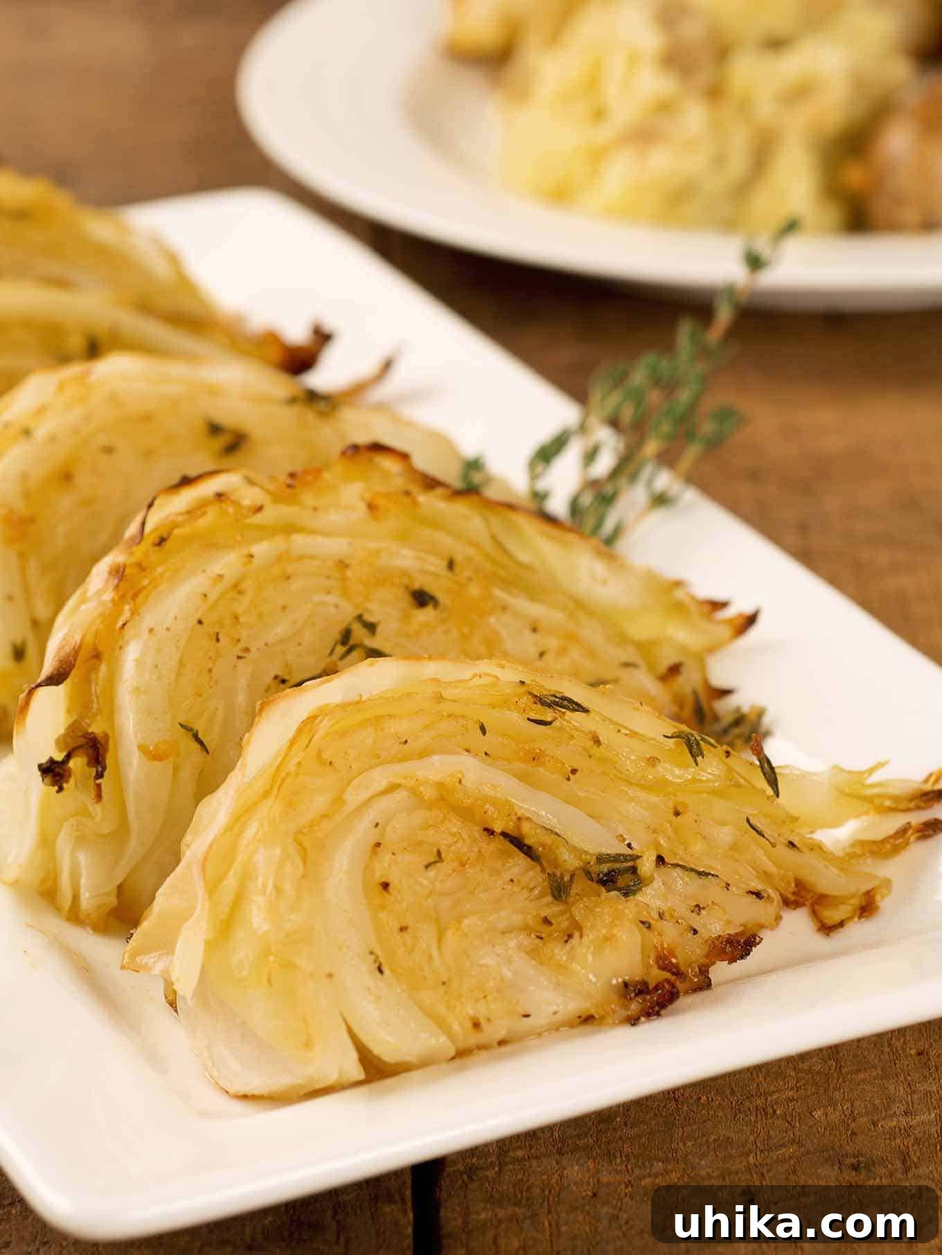 Aromatic Garlic Herb Roasted Cabbage 2 Wedges of golden, herb and garlic roasted cabbage on a white serving plate, garnished with fresh thyme sprigs. A dinner plate is visible in the soft-focus background, suggesting a complete meal.