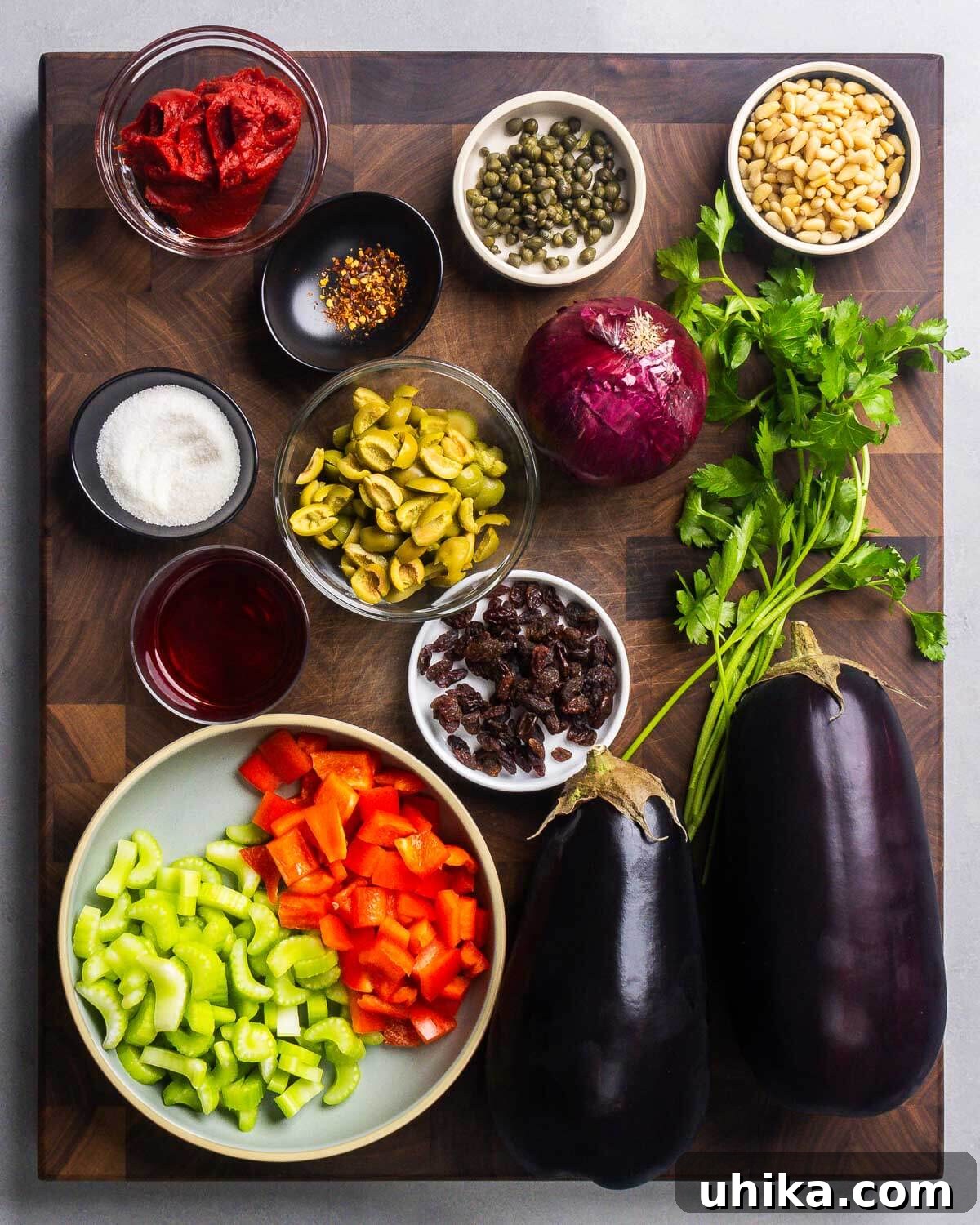 A flat lay photograph showcasing all the fresh and pantry ingredients needed for making eggplant caponata, including eggplant, celery, bell pepper, onion, tomato paste, olives, capers, raisins, pine nuts, and fresh parsley.