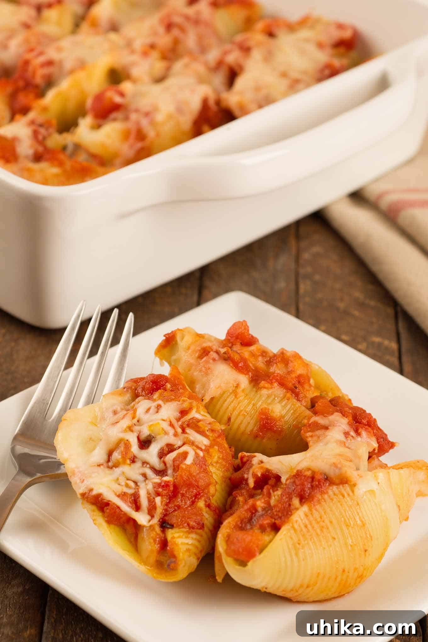 Savory Stuffed Sausage Shells 2 Sausage stuffed pasta shells topped with marinara and melted cheese, served on a white plate with a fork, the casserole with more shells in the background. A delightful Italian-American comfort food dish perfect for family dinners.