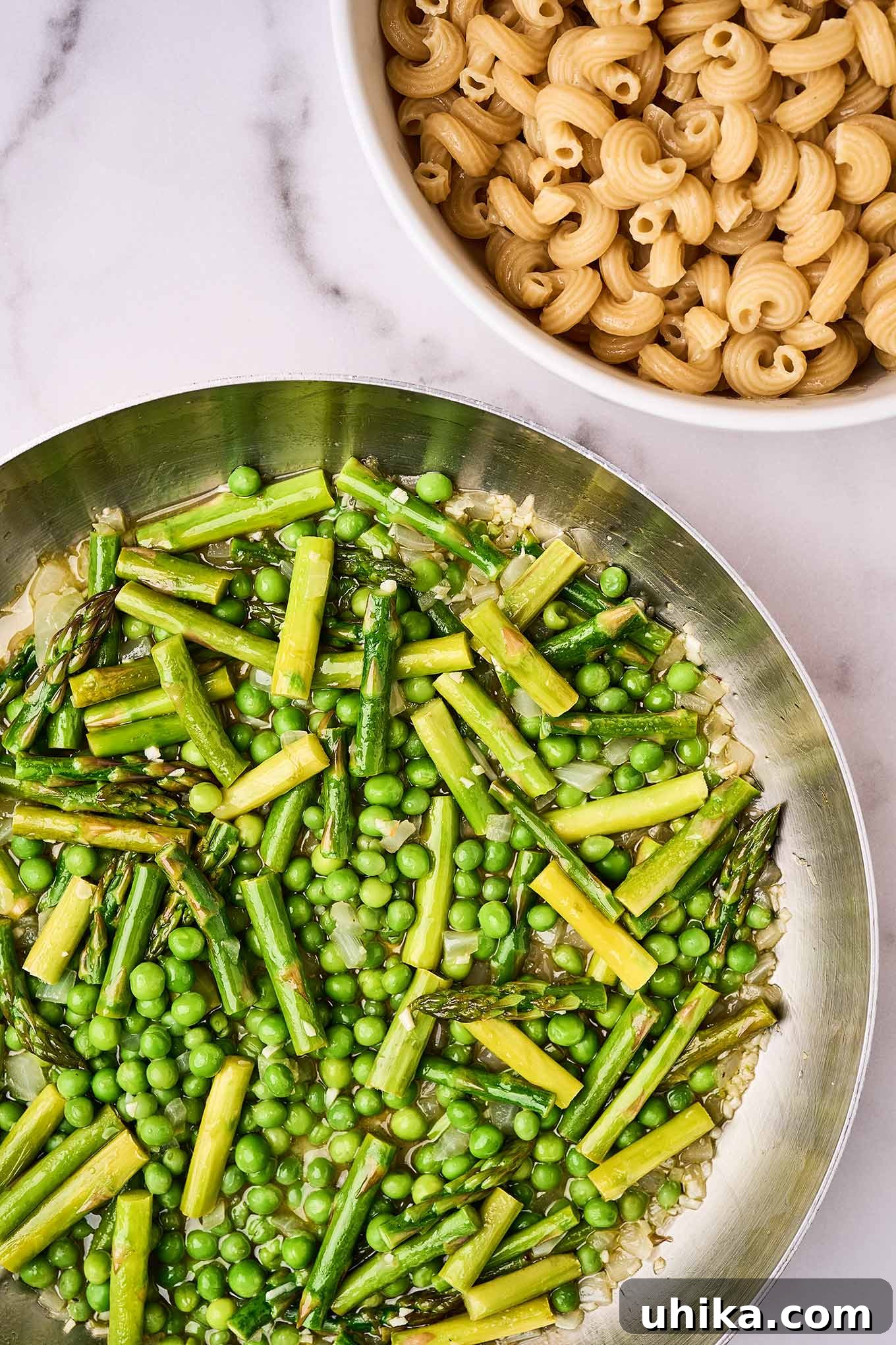 Vibrant Shrimp Primavera 5 A frying pan with sautéed garlic, onion, asparagus, and sweet peas. Uncooked cavatappi pasta sits in a bowl nearby.