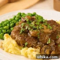 Salisbury steak topped with mushroom and onion gravy, served with mashed potatoes and peas on a white plate.