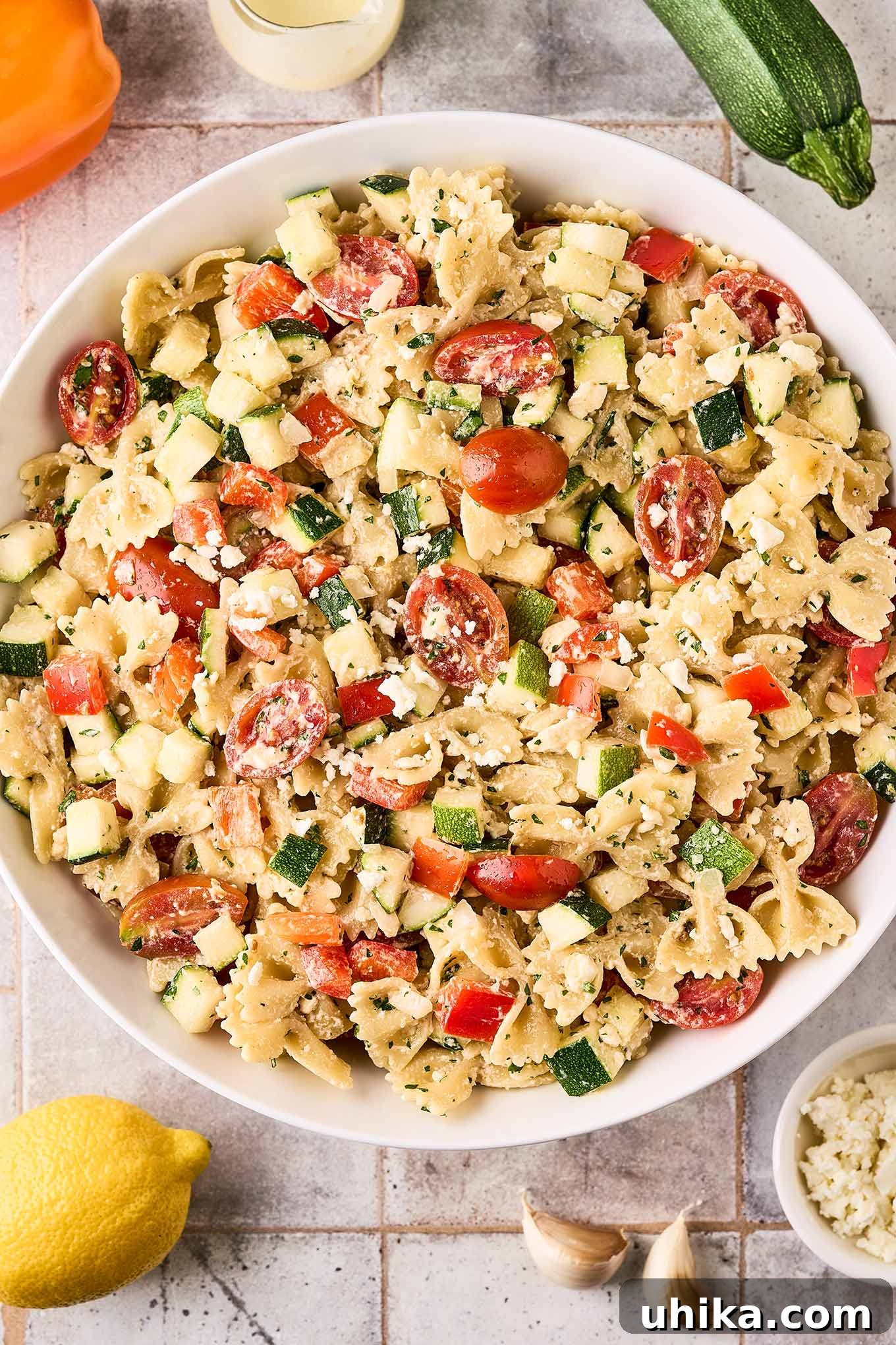 Zesty Lemon Cream Pasta Salad 2 An overhead view of a serving bowl of creamy pasta salad with lemon dressing on a tile surface surrounded by recipe ingredients.