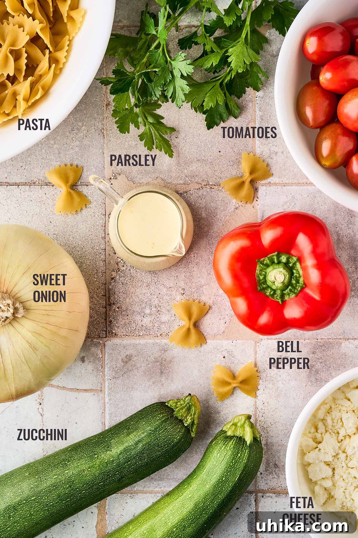 Zesty Lemon Cream Pasta Salad 3 A labeled, overhead view of the ingredients for creamy pasta salad; pasta, parsley, tomatoes, onion, red bell pepper, zucchini, and feta cheese.