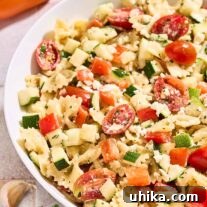 A bowl of creamy pasta salad with lemon dressing surrounded by an orange bell pepper, a dish of crumbled feta, garlic cloves, and a sprig of parsley.