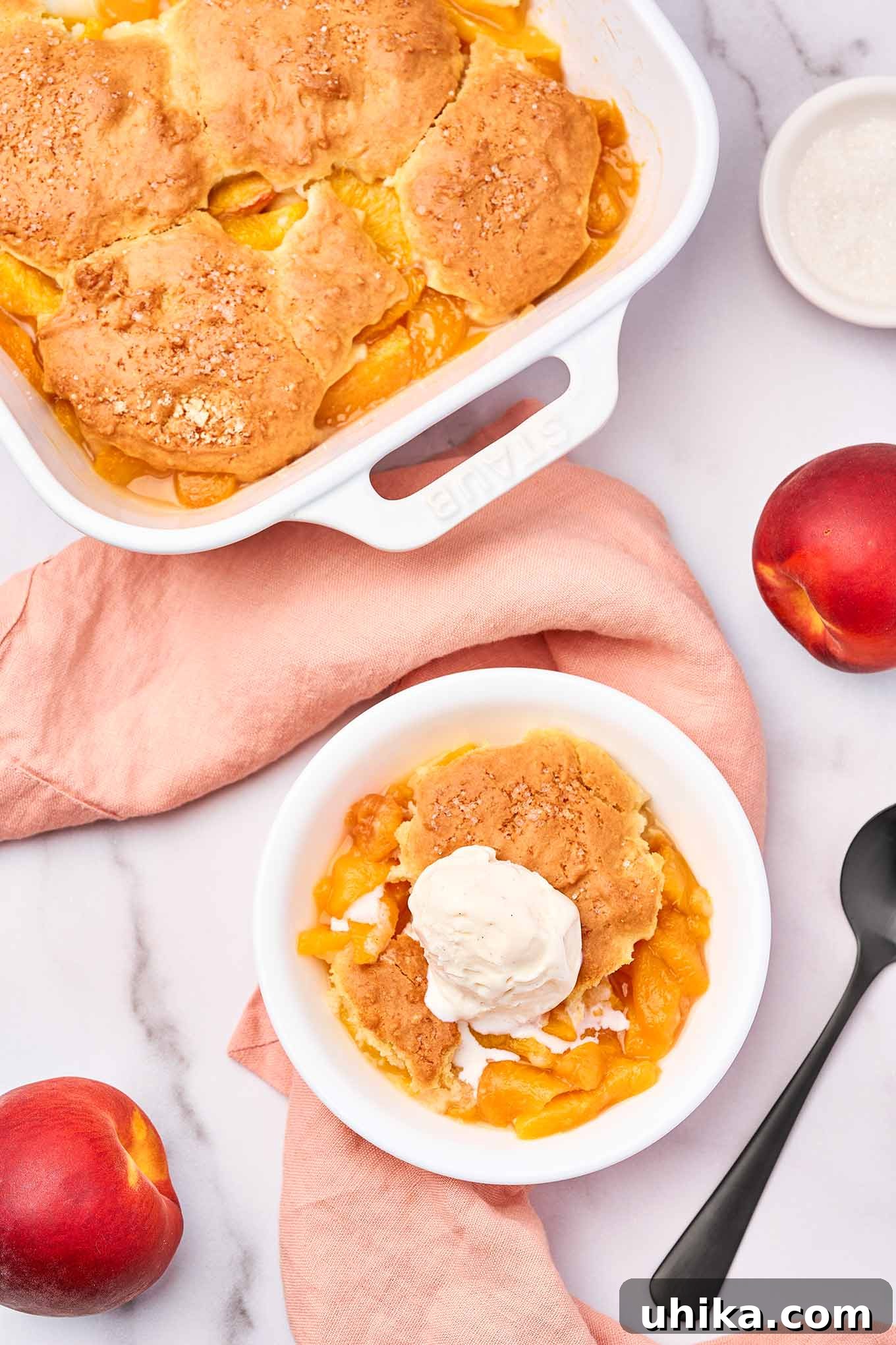Summertime Fresh Peach Cobbler 4 A dessert bowl of peach cobbler topped with vanilla ice cream next to a spoon, whole peaches and the cobbler casserole to the side.