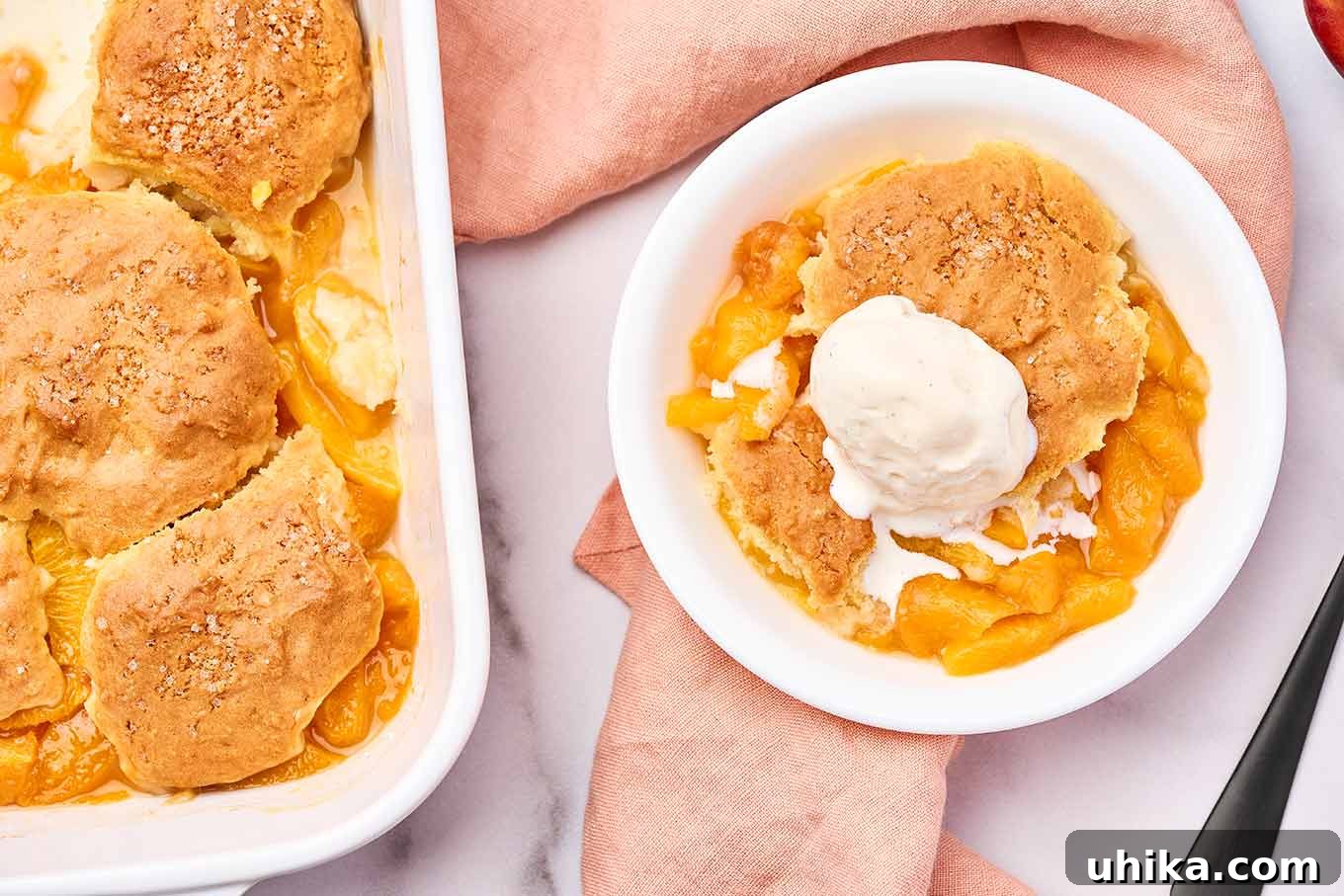 Summertime Fresh Peach Cobbler 7 An overhead view of a white baking dish filled with peach cobbler next to a single serving of the dessert topped with ice cream.