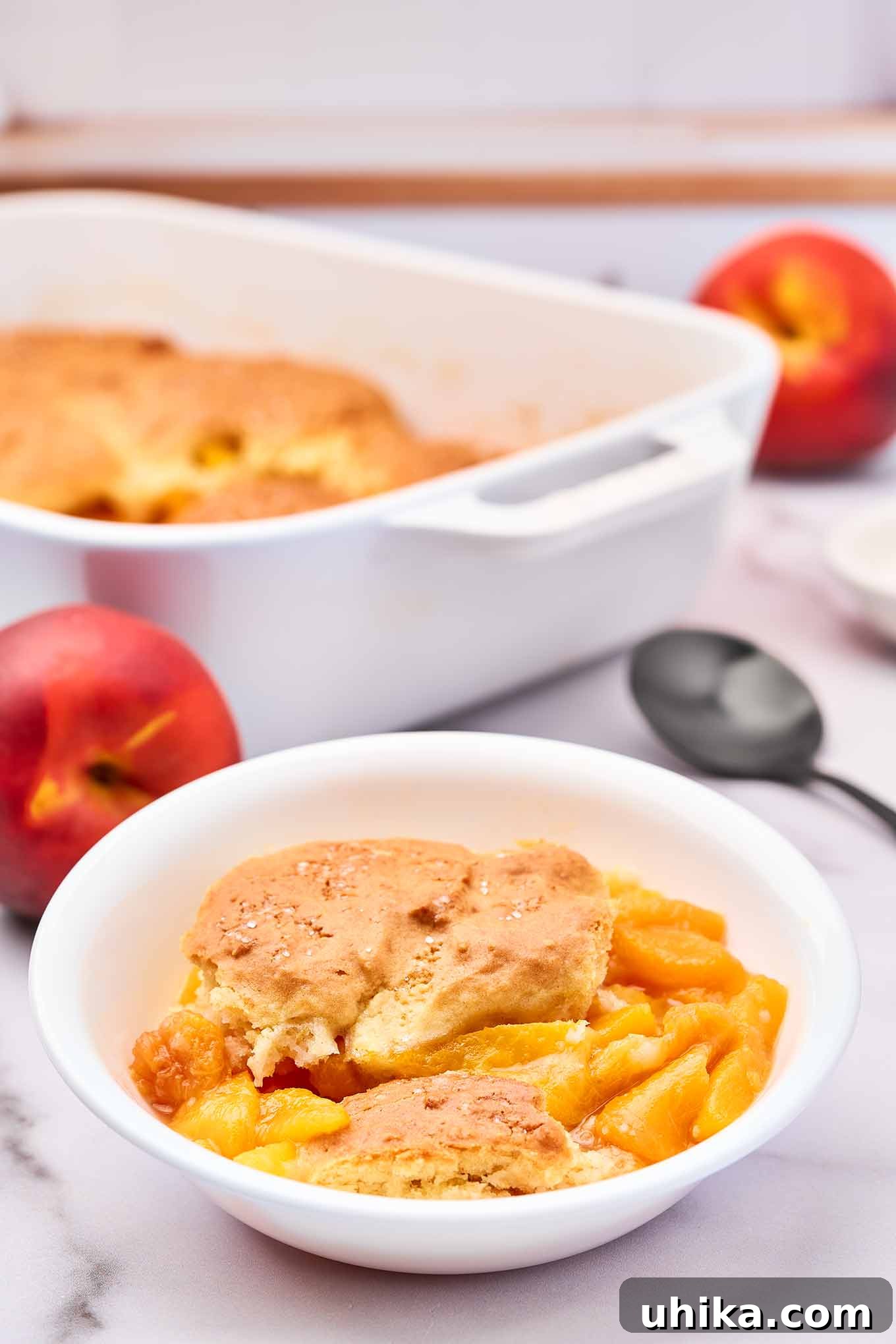 Summertime Fresh Peach Cobbler 8 A single serving of warm peach cobbler in a white dessert bowl with the whole cobbler and a spoon in the background.