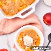 A dessert bowl of peach cobbler topped with vanilla ice cream next to a spoon, whole peaches and the cobbler casserole to the side.