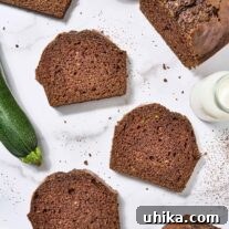 A partially sliced loaf of zucchini bread on a marble surface surrounded by slices of bread, a prep bowl of shredded zucchini, a whole zucchini, cup of flour, and pitcher of buttermilk.
