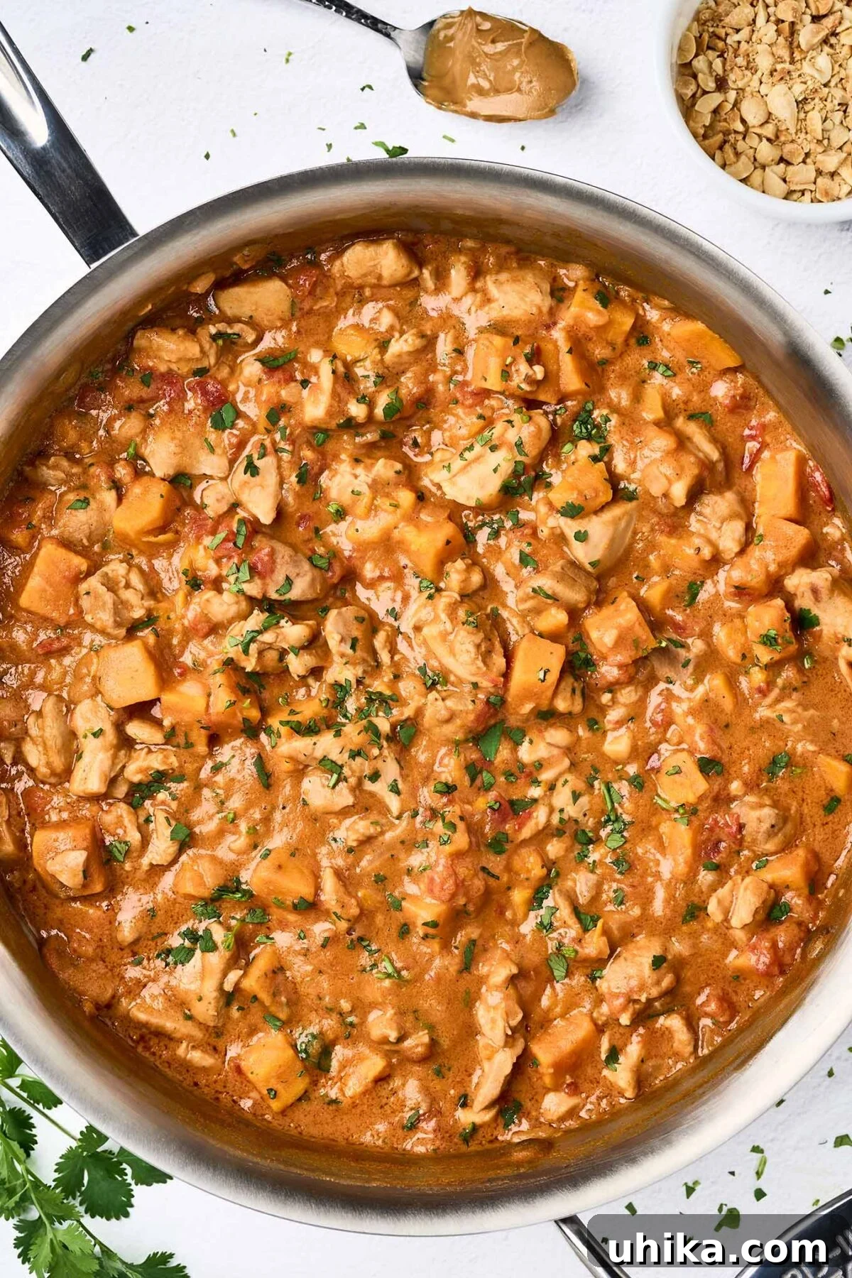 Velvety African Peanut Chicken Pot 5 A large sauté pan brimming with rich, bubbling African peanut stew, showcasing tender chunks of chicken thighs and vibrant sweet potato cubes submerged in a thick, savory, peanut butter and tomato-based sauce, ready to be served.