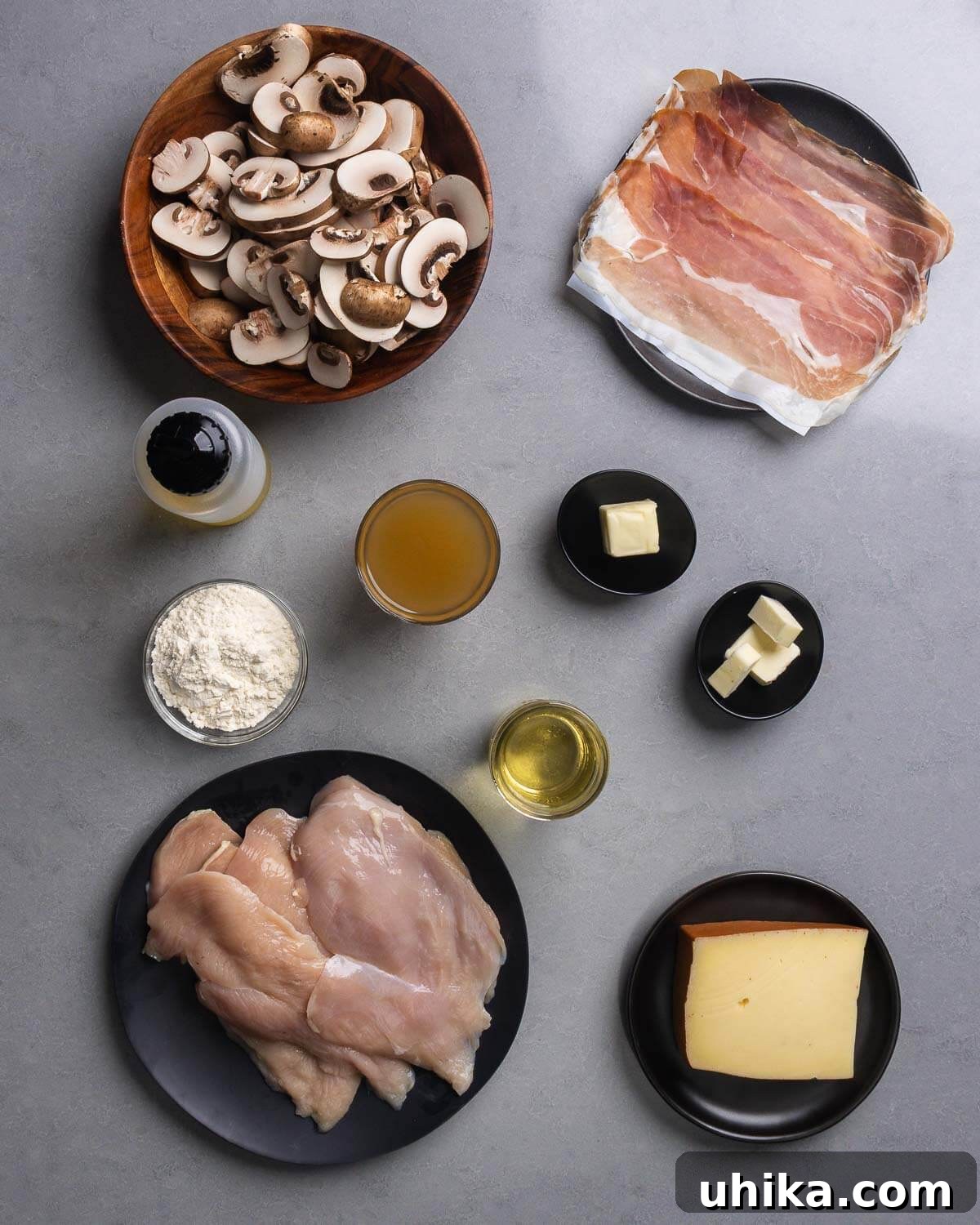 Classic Chicken Valdostana 4 A meticulously arranged flat lay of all the fresh ingredients required for Chicken Valdostana, including sliced cremini mushrooms, delicate prosciutto, a bottle of olive oil, a bowl of all-purpose flour, a carton of chicken stock, a stick of butter, a bottle of dry white wine, raw chicken cutlets, and a block of Fontina cheese.