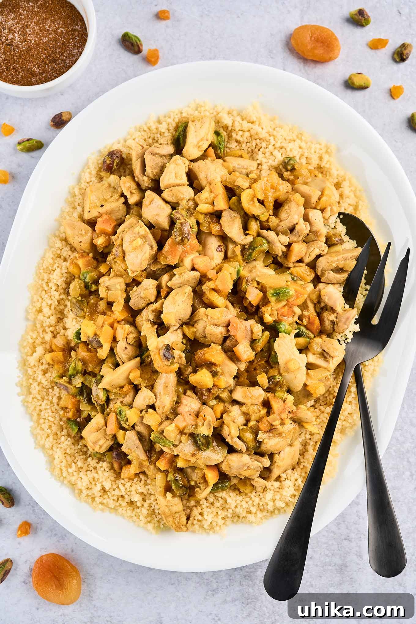 Jeweled Apricot Pistachio Chicken 2 An oval serving platter of couscous topped with Persian style apricot chicken and pistachios with serving utensils, surrounded by a prep bowl of the spice blend, dried apricots, and chopped pistachios.