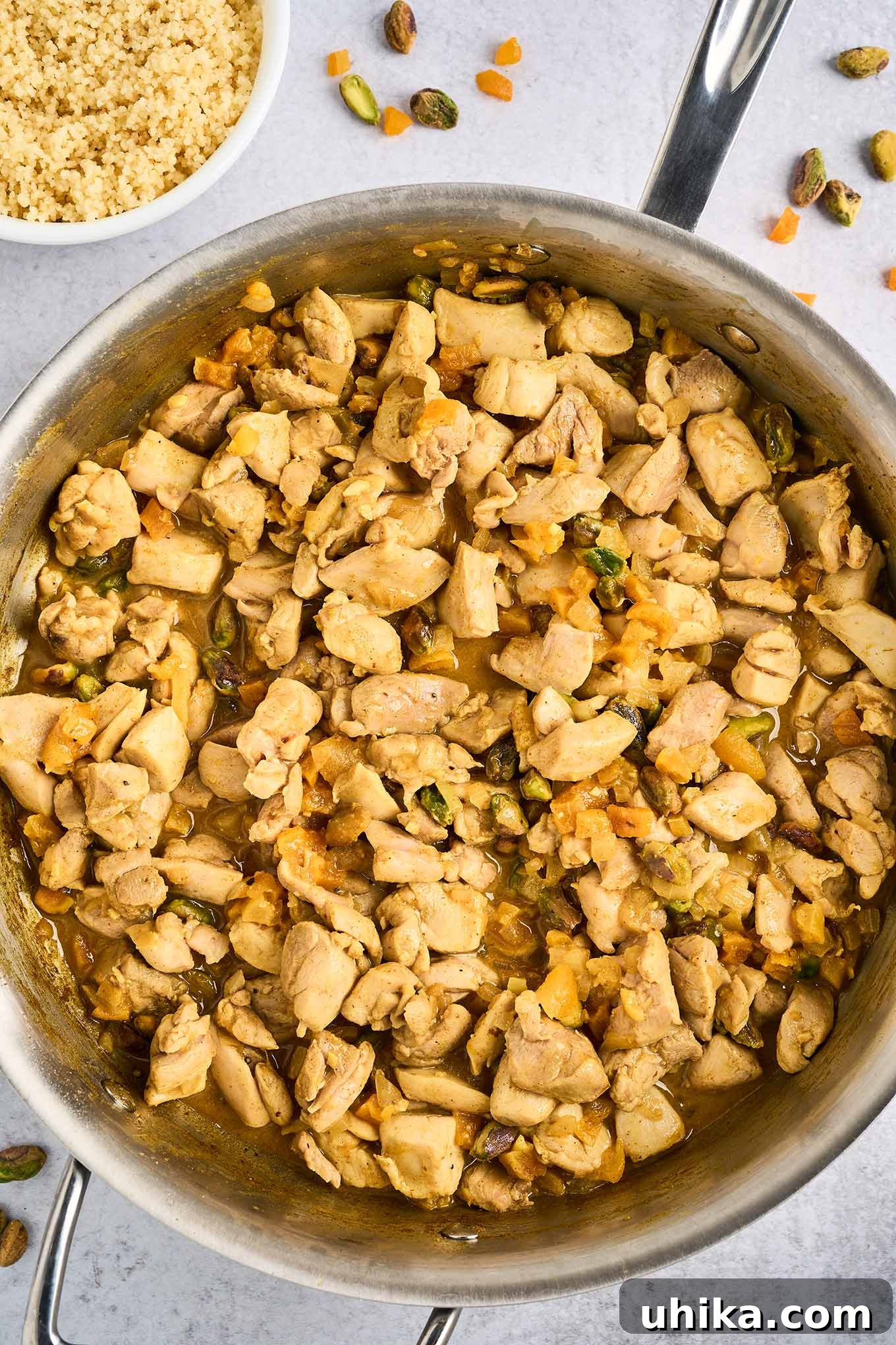 Jeweled Apricot Pistachio Chicken 5 A frying pan filled with chunks of tender chicken thighs combined with spices, dried apricots, pistachio nuts, sautéed garlic and onion, lemon juice and zest, and butter, a bowl of couscous in the background.