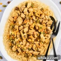 An oval serving platter of couscous topped with Persian style apricot chicken and pistachios with serving utensils, surrounded by a prep bowl of the spice blend, dried apricots, and chopped pistachios.
