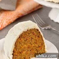 A slice of moist, spiced carrot cake with cream cheese frosting on a plate, the remainder of a cake on a stand in the background along with a fork and printed cloth napkin.