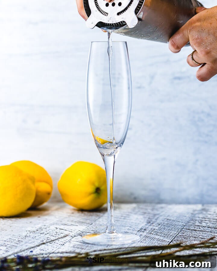 Pouring Lavender French 75 into Flute Hand pouring a Lavender French 75 cocktail into a champagne flute, with three lemons in the soft background.