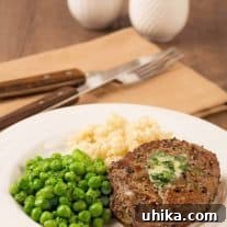 A peppercorn crusted filet mignon topped with compound butter on a plate with mashed potatoes and peas, napkin with fork and knife in the background.