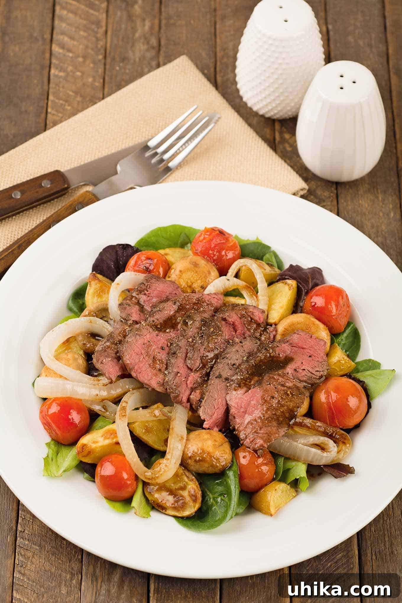 Robust Steak and Roasted Potato Salad 2 A vibrant plate featuring a warm salad of perfectly grilled sirloin steak slices, golden baby potatoes, bursting cherry tomatoes, and savory mushrooms, all nestled over a bed of fresh mixed greens. A clean napkin with a fork and knife rests beside the plate, inviting a delicious meal.