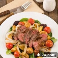 A plate with a salad of grilled sirloin, baby potatoes, cherry tomatoes and mushrooms over mixed greens, napkin with fork and knife to the side.