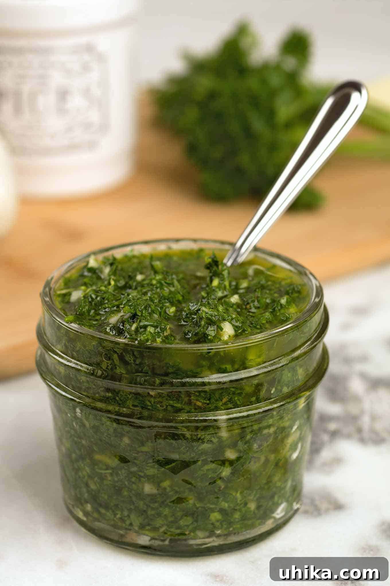 Zesty Homemade Chimichurri 2 A quilted glass jam jar of chimichurri sauce (parsley, cilantro, garlic, olive oil, red wine vinegar, and seasoning) with a spoon, a cutting board with fresh herbs and seasonings in the background.