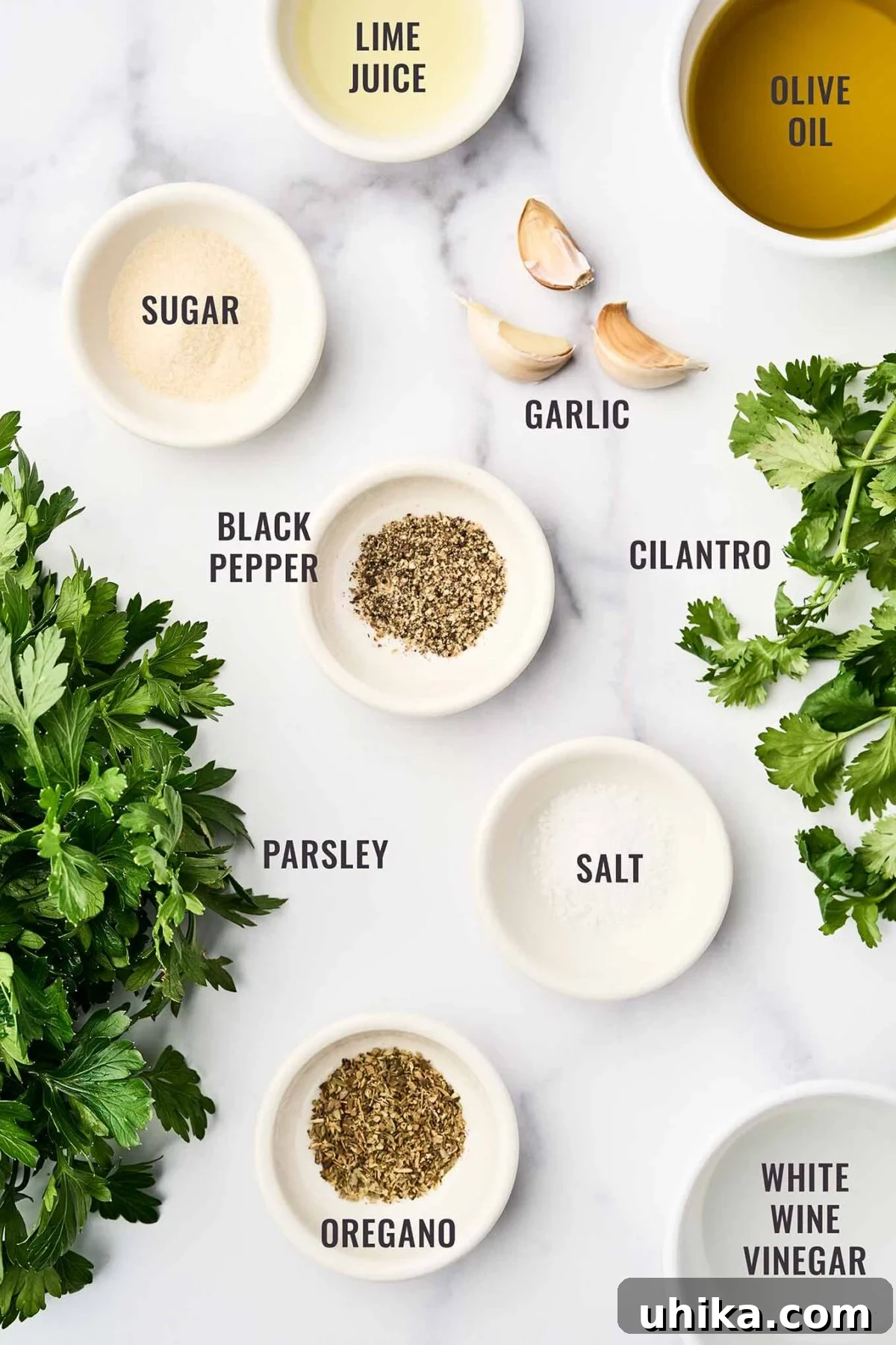 Zesty Homemade Chimichurri 3 A labeled, overhead view of the ingredients for chimichurri sauce including parsley, cilantro, garlic, oregano, black pepper, salt, sugar, lime juice, white wine vinegar, and olive oil.