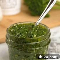 Zesty Homemade Chimichurri 5 A quilted glass jam jar of chimichurri sauce (parsley, cilantro, garlic, olive oil, red wine vinegar, and seasoning) with a spoon, a cutting board with fresh herbs and seasonings in the background.