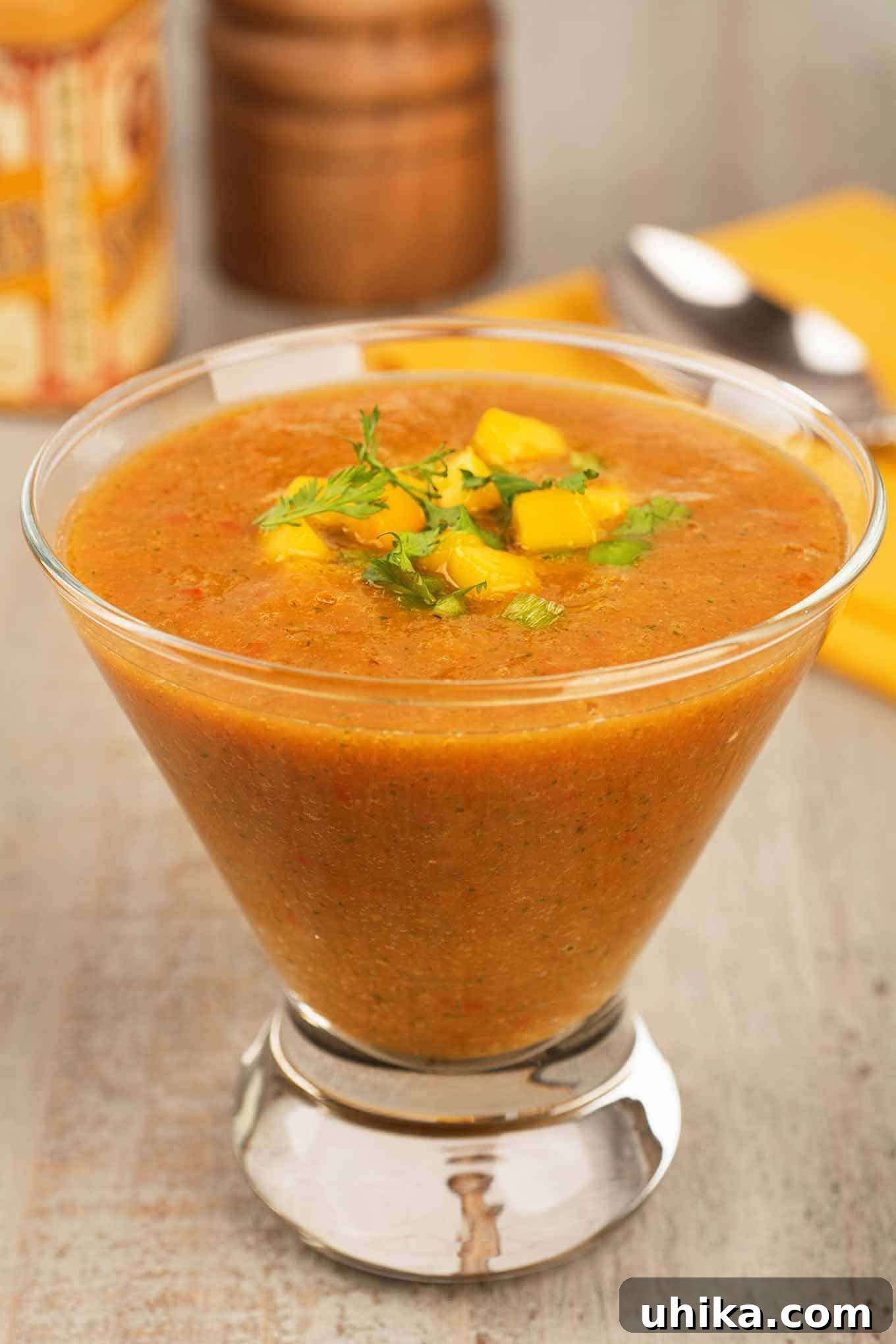 Sunshine Mango Gazpacho 2 A refreshing serving of sweet and savory mango gazpacho soup, garnished with vibrant chopped mango and fresh cilantro, presented in a clear glass alongside a napkin, spoon, pepper mill, and seasoning jar.