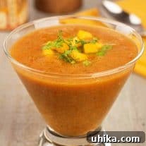 A serving of mango gazpacho soup, garnished with chopped mango and cilantro, served in a clear glass with a napkin, spoon, pepper mill, and jar of seasoning in the background.