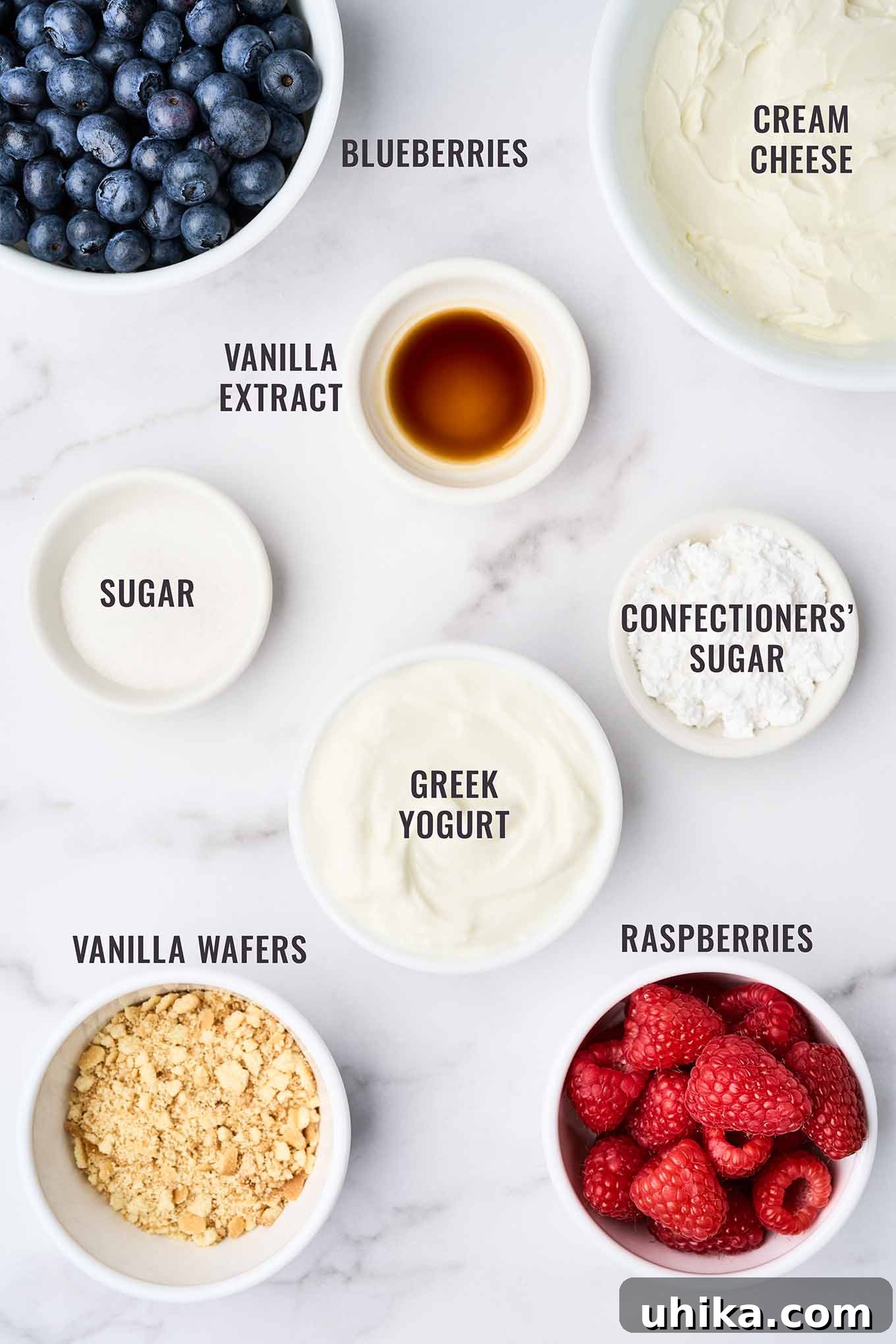 Layered No-Bake Cheesecake Delights 3 An overhead view of the ingredients for no-bake cheesecake parfaits with fresh berries; cream cheese, Greek vanilla yogurt, confectioners' sugar, vanilla wafers, raspberries, blueberries, and vanilla extract.