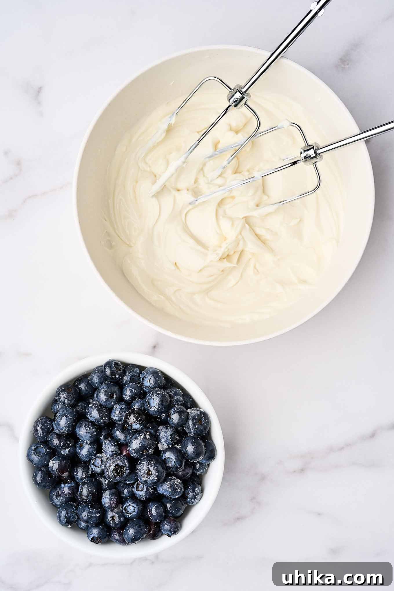 Layered No-Bake Cheesecake Delights 4 Cheesecake parfait preparation; whipped cream cheese and Greek vanilla yogurt in a mixing bowl with electric mixer beaters beside a bowl of sugared blueberries.