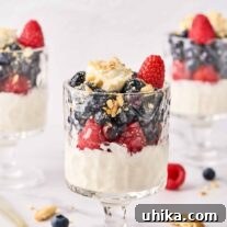 Three cheesecake parfaits, each topped with blueberries, raspberries, and crumbled vanilla cookies in footed parfait glasses, surrounded by cookie crumbs, whole berries, and a spoon.
