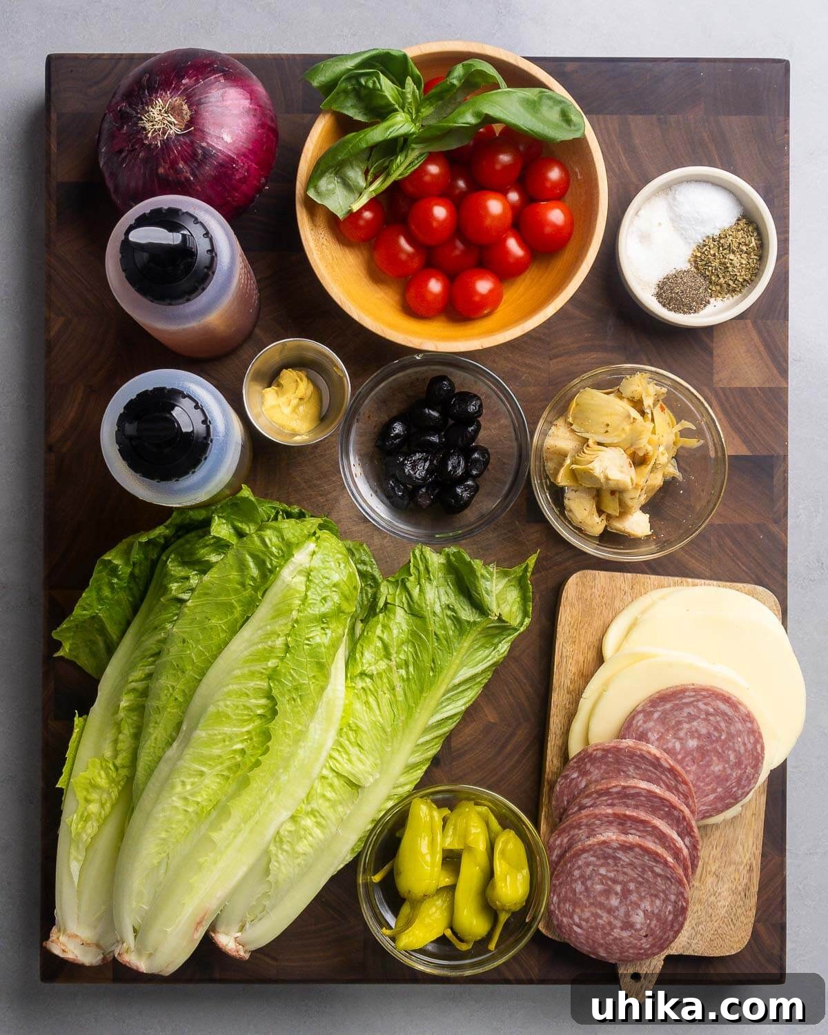 Italian Antipasto Chopped Salad 3 A colorful array of ingredients including red onion, fresh basil, cherry tomatoes, various spices, red wine vinegar, olive oil, Dijon mustard, olives, artichoke hearts, crisp lettuce, pepperoncini, provolone cheese, and Genoa salami, laid out on a wooden board.
