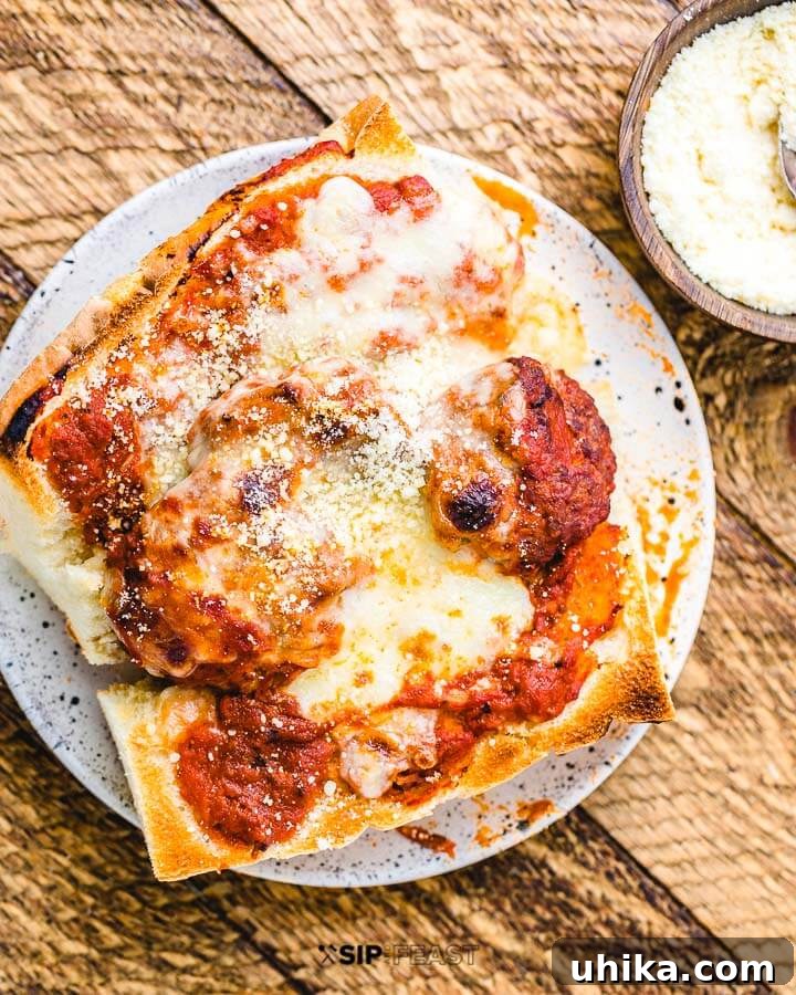 Meatball Parm Hero on Plate A single, delectable meatball parm sub, cut in half, resting on a white plate with a side of grated Parmesan cheese, on a rustic wooden table.