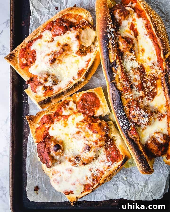 Baked Meatball Parm Heroes Three perfectly baked meatball parm heroes, golden and bubbly with melted cheese, resting on a parchment-lined sheet pan, fresh from the oven.