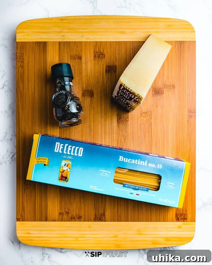 Peppery Cheese Bucatini 2 Ingredients for Cacio e pepe: A block of Locatelli Pecorino Romano cheese, a black pepper grinder, and uncooked bucatini pasta elegantly displayed on a wooden cutting board.