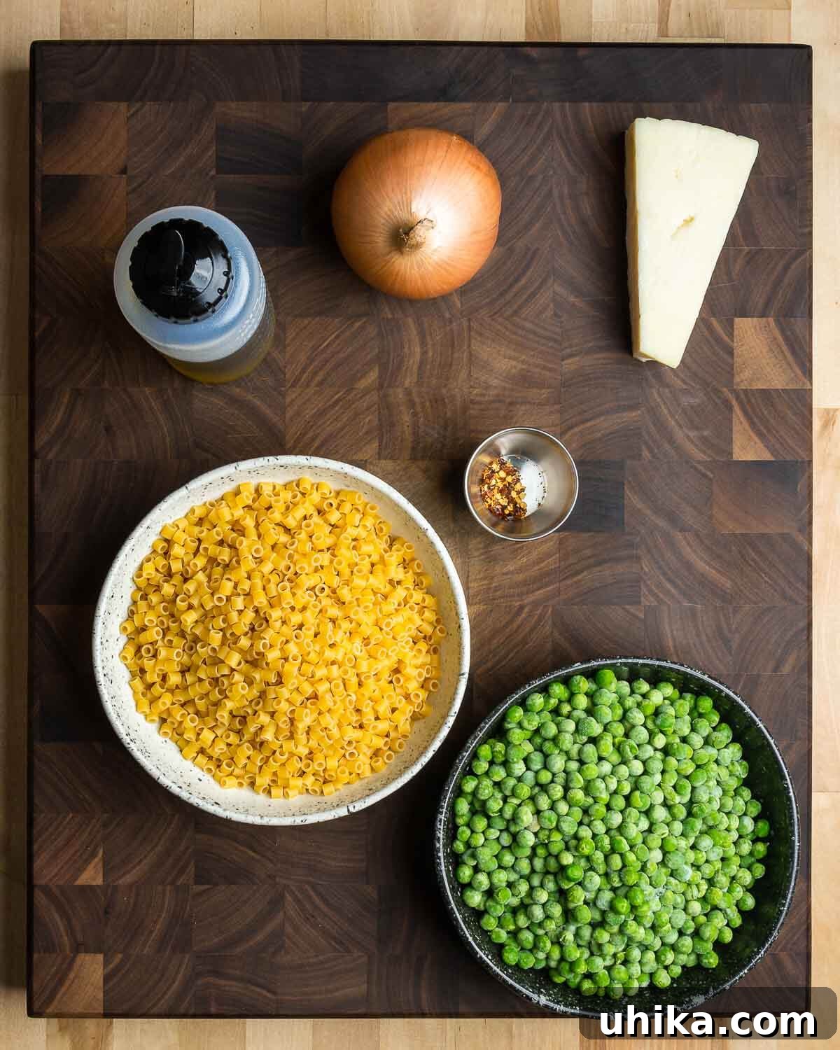 Sweet Pea Pasta 3 A flat lay photograph showcasing all the ingredients required for Pasta e Piselli: high-quality olive oil, a diced onion, freshly grated Pecorino Romano cheese, ditalini pasta, a pinch of hot red pepper flakes, and a bag of frozen peas.
