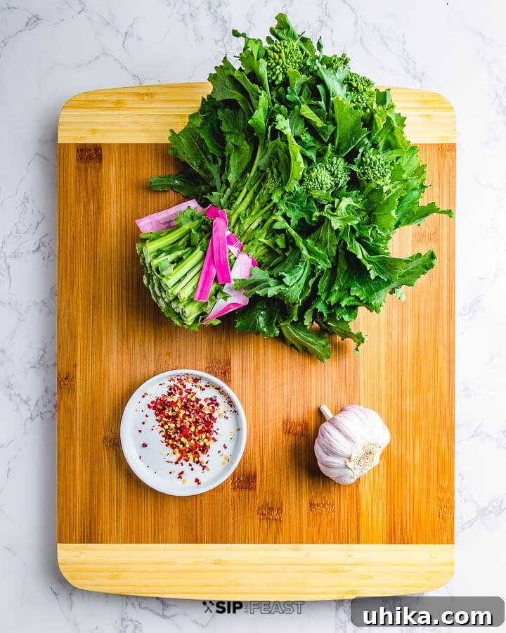 Broccoli Rabe Saute Ingredients - Sip & Feast #broccolirabeingredients #garlicrecipes #spicygreens Fresh ingredients laid out on a cutting board: a vibrant bunch of broccoli rabe, whole garlic cloves, and a small pile of red pepper flakes.