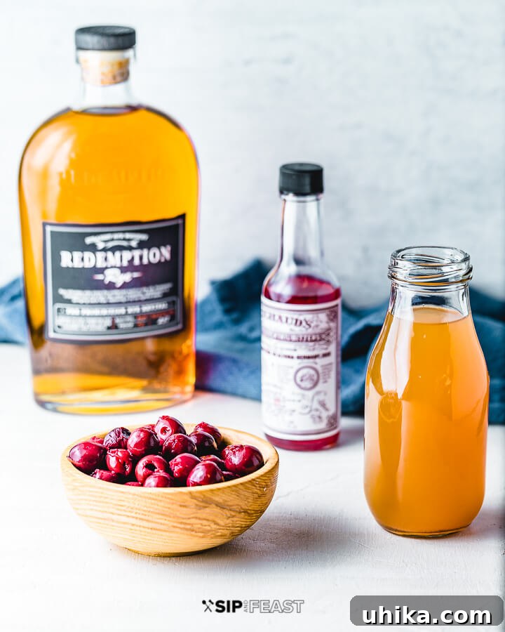 A bottle of rye whiskey, cocktail bitters, a bowl of fresh cherries, and a jug of apple cider, all essential ingredients for the Apple Cider Old Fashioned.