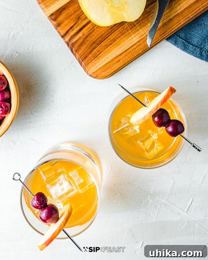Two Apple Cider Old Fashioned cocktails viewed from above, with a cutting board and a half-eaten apple in the top right corner, showcasing the drink's presentation.