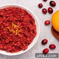 A serving bowl of fresh cranberry orange relish garnished with orange zest, on a gray background with scattered cranberries to the right.