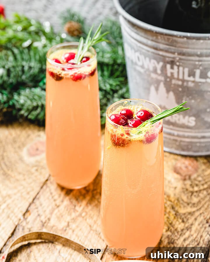 Two champagne flutes filled with vibrant cranberry mimosas, garnished with fresh cranberries and a sprig of rosemary. A bucket with a bottle of Prosecco chills in the soft-focus background, suggesting a festive atmosphere.